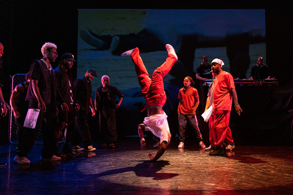 in a circle formation the company of dancers some dressed in black the others in red look on as a dancer in red pants seems suspended upside down in mid-air
